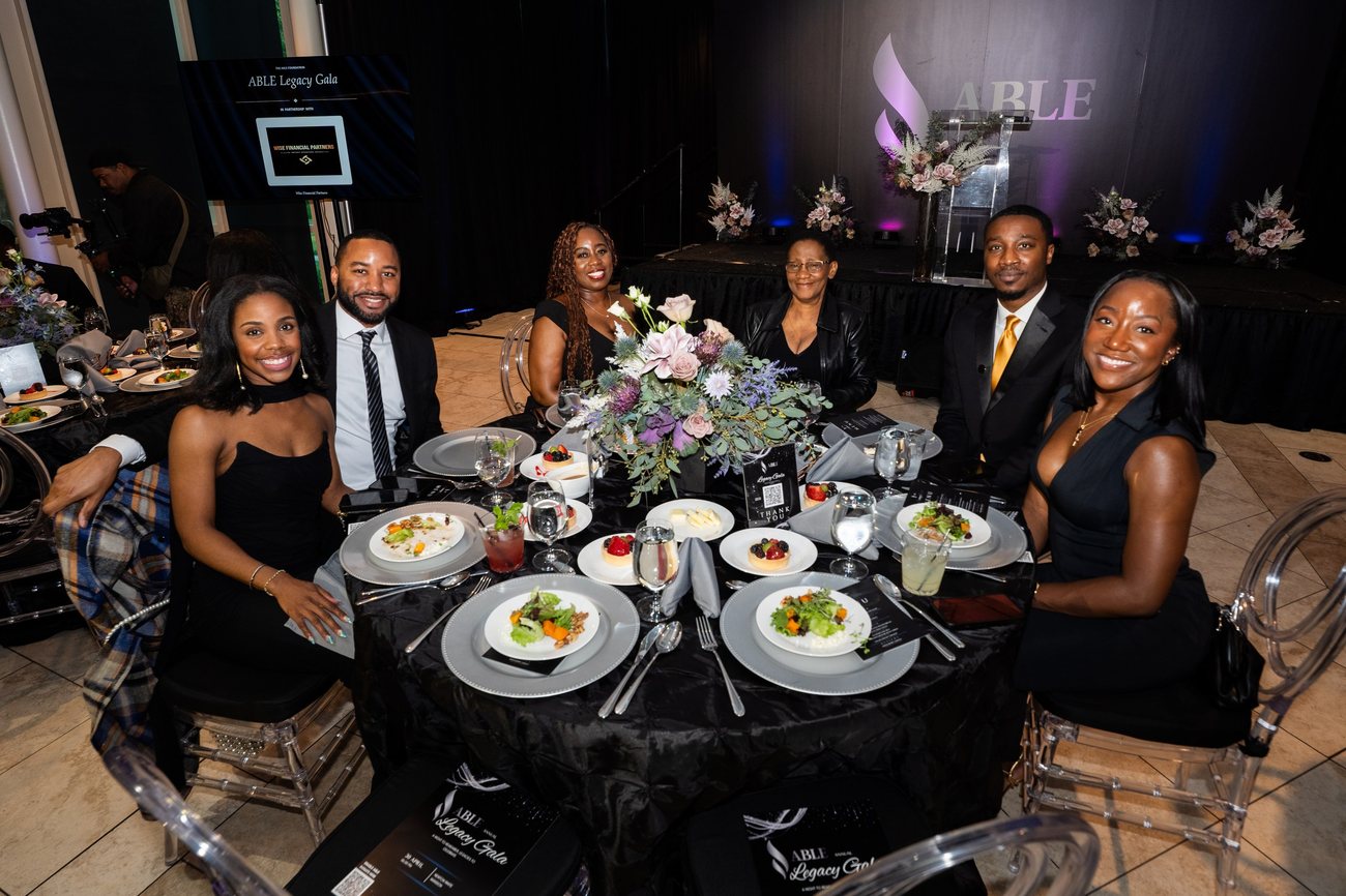 Six guests smiling around a dinner table at the ABLE Legacy Gala, the stage behind them